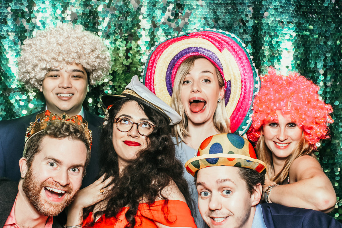 group of guests at a wedding posing with wigs and hats in front of a green sequins backdrop at Elmore Court wedding venue