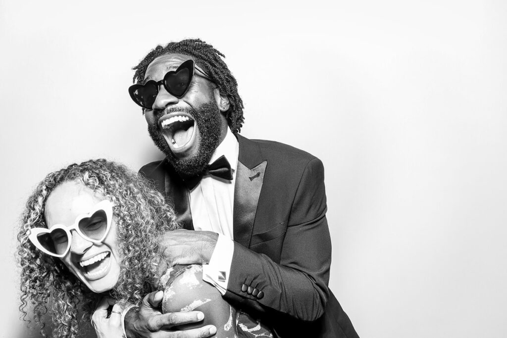 A couple bursting into laughter, enjoying the timeless look of the black and white photo booth setup during a wedding party entertainment at Elmore Court in Gloucester