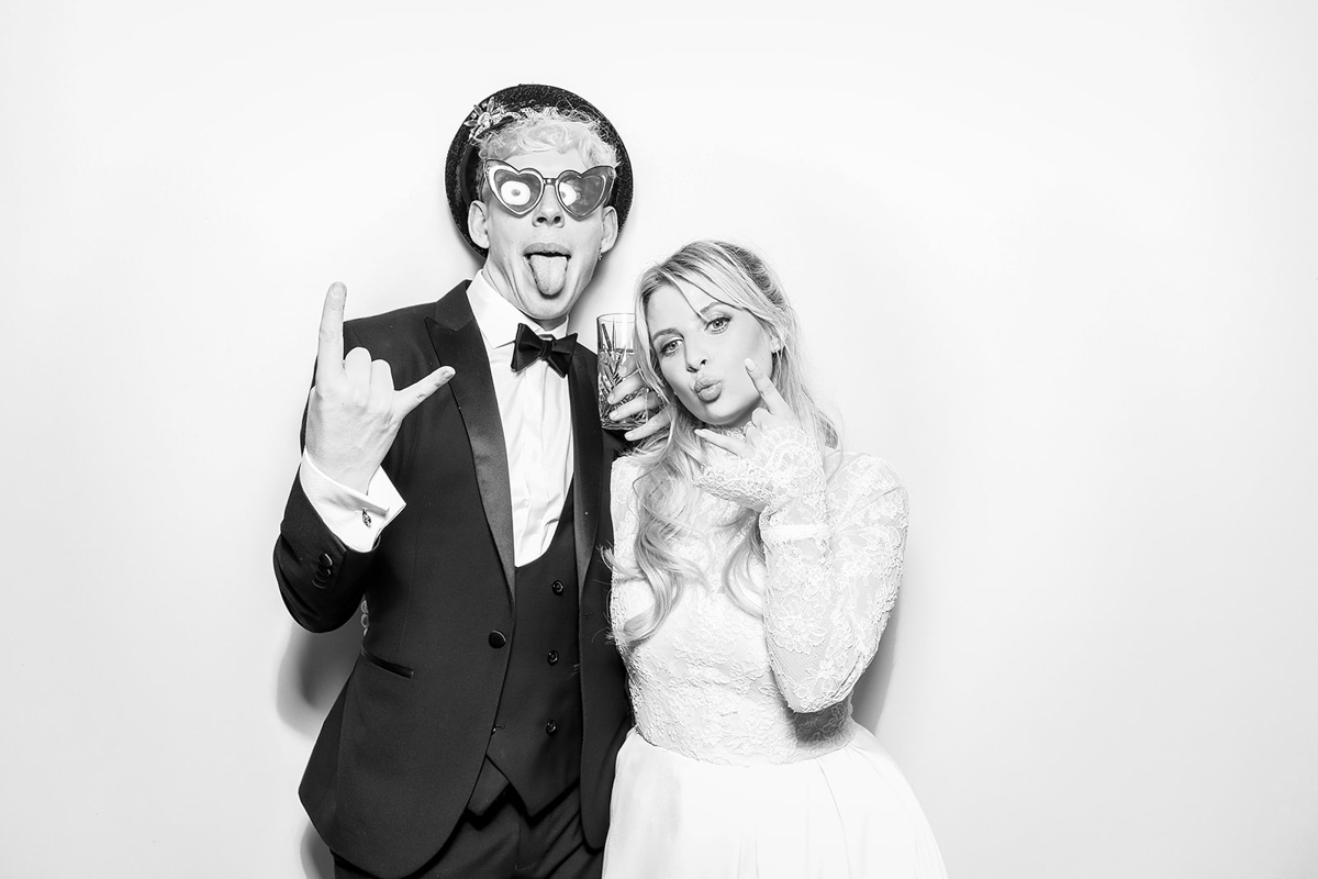 A groom in a tuxedo and bride in a lace gown posing playfully in a black and white Kardashian-style photo booth.