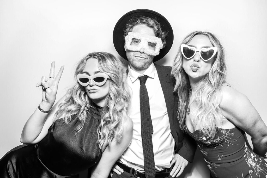 Three elegantly dressed guests posing in a photo booth wearing quirky sunglasses and accessories during the wedding party entertainment at a Gloucester wedding venue, using the B&W filter for the photo booth