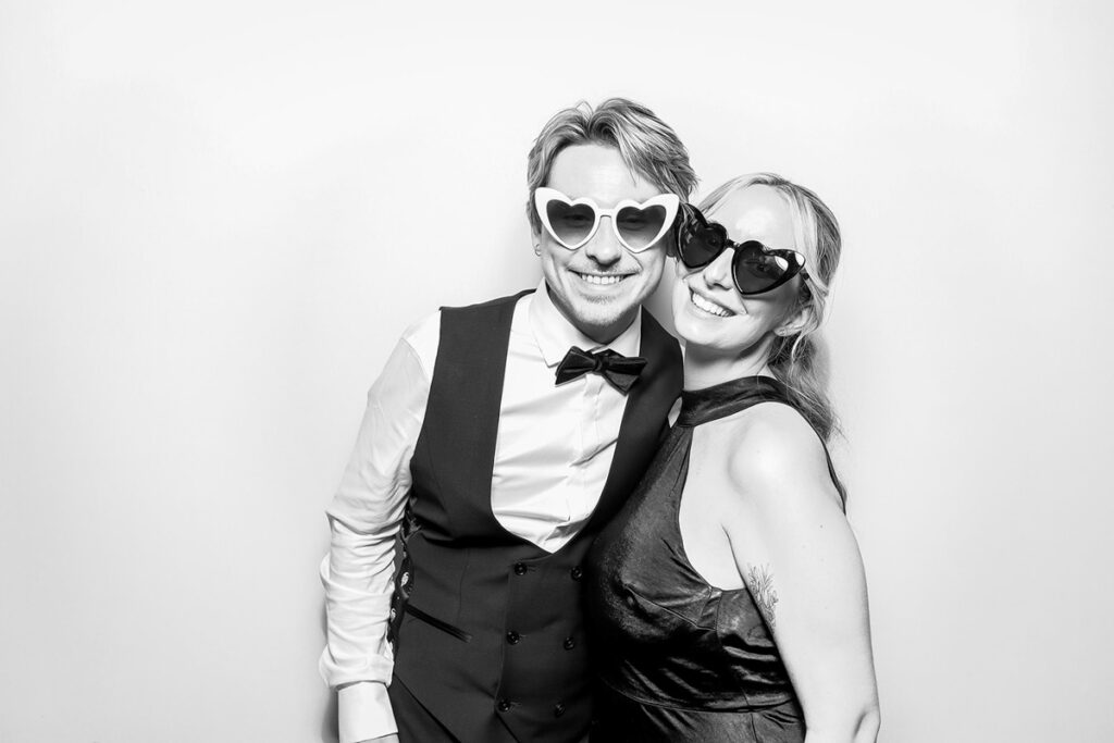 A well-dressed couple smiling and posing in a b& white photo booth filter with heart-shaped sunglasses at Elmore Court wedding venue, gloucester