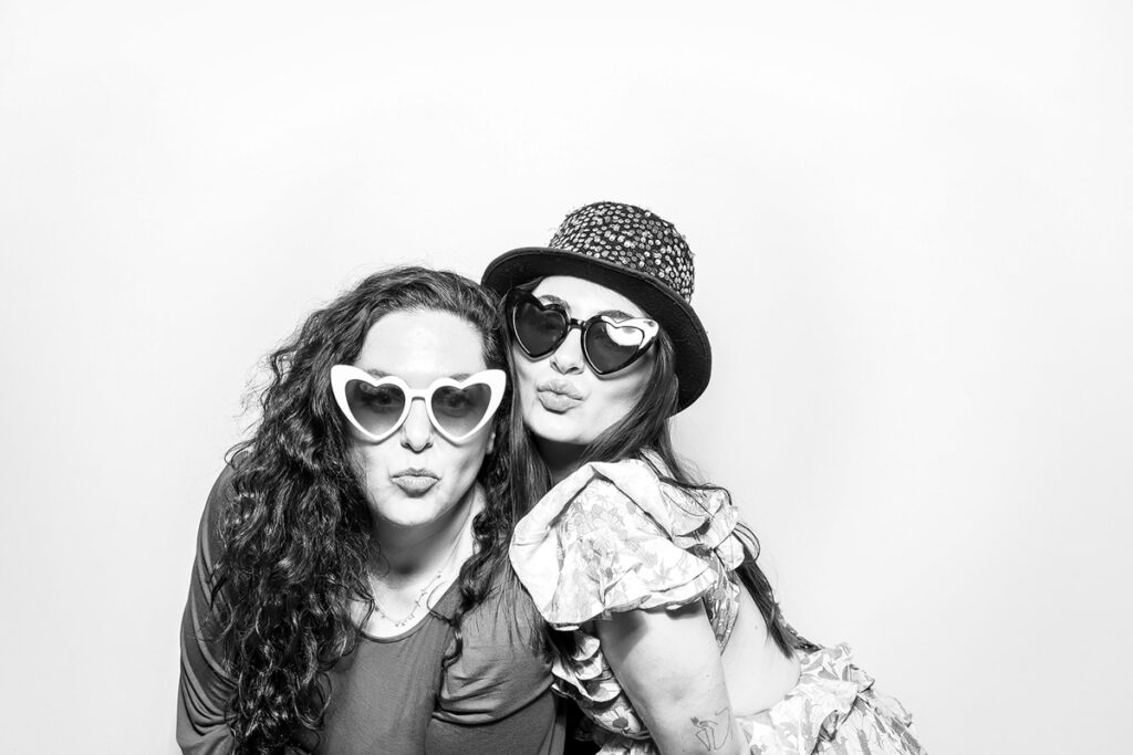 Two guests wearing heart-shaped sunglasses, striking a playful pose in a black and white Kardashian-style photo booth.