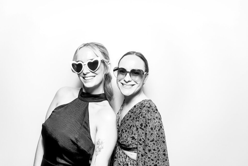 Smiling friends dressed elegantly, posing with heart-shaped sunglasses against a sleek black and white photo booth backdrop