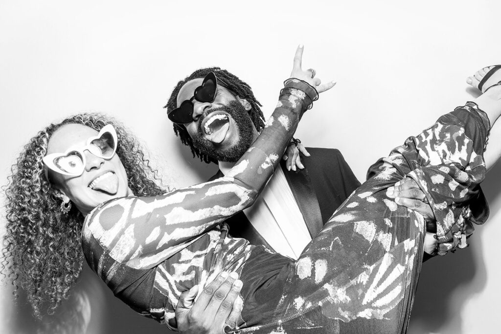 A fun lift pose by a cheerful couple, wearing quirky sunglasses and captured in a stylish black and white booth filter.