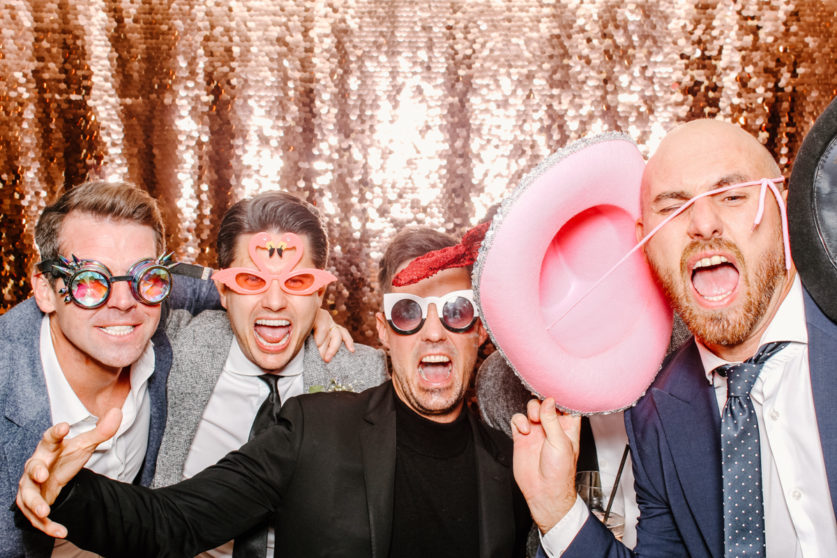 guys posing in a fun way during a wedding party entertainment, wearing fun props and with a sequins backdrop behind 