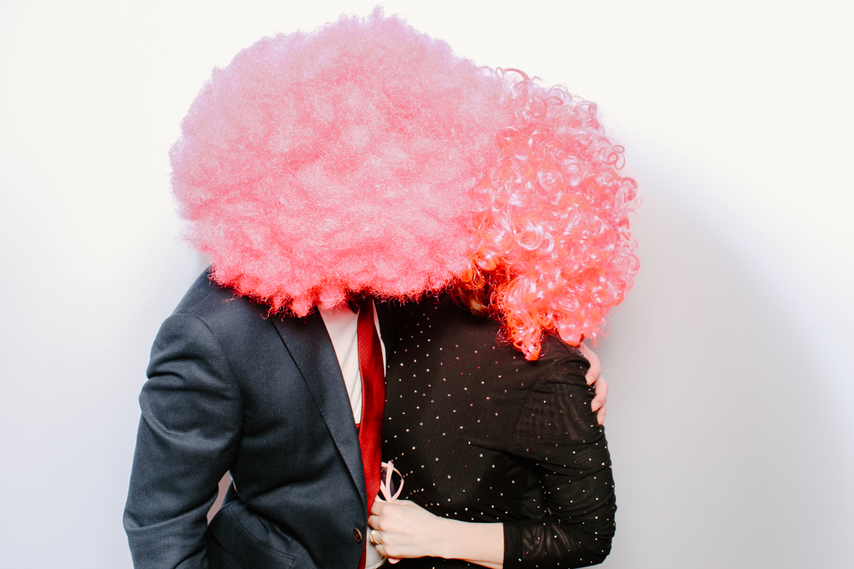 couple with large pink wigs for a wedding party photo booth entertainment with white backdrop