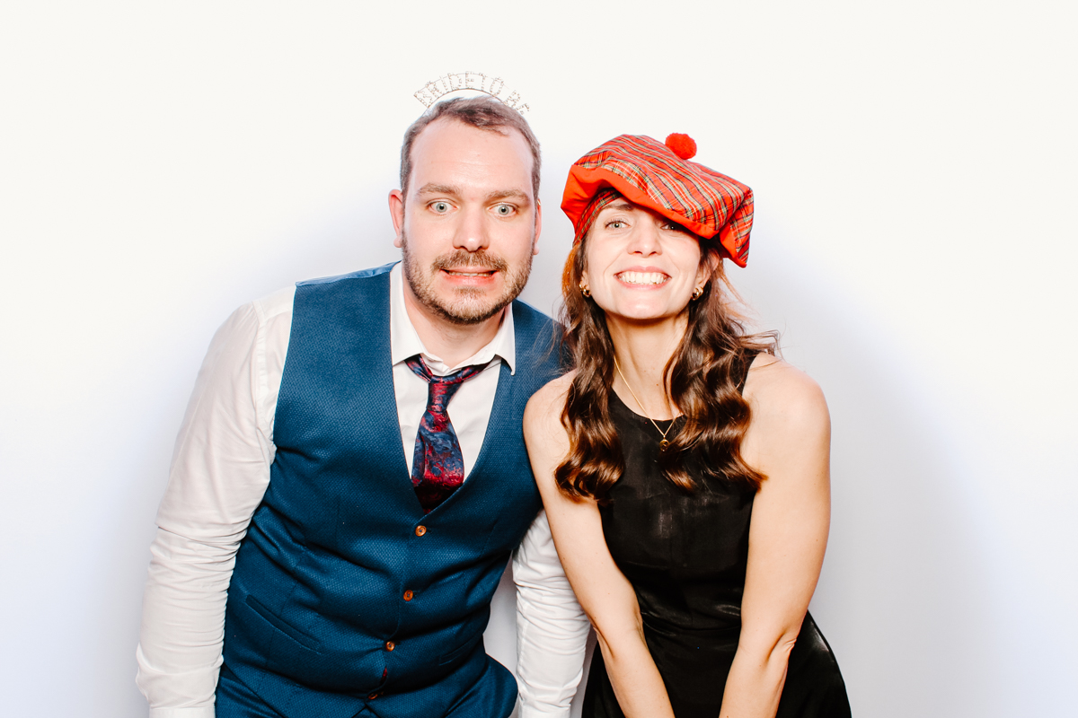 couple posing for mad hat photo booth during a wedding party with white backdrop