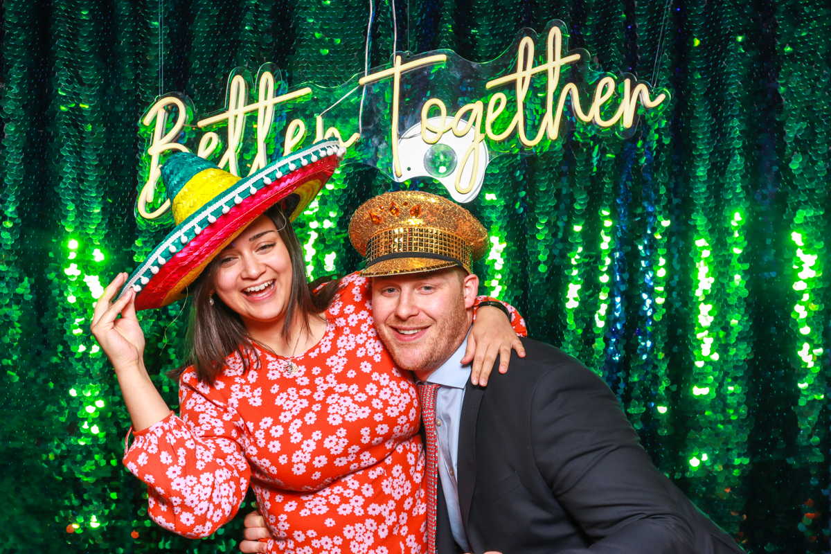 couple posing with neon sign and green sequins backdrop for mad hat photo booth
