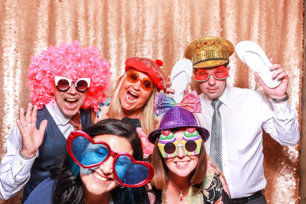 5 guests posing with props, against a blush sequins backdrop, for a Wedding Photo Booth rental