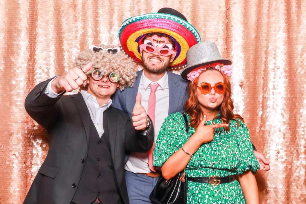 3 guests posing with props, against a blush sequins backdrop, for a Bredenbury Court Wedding Photo Booth rental