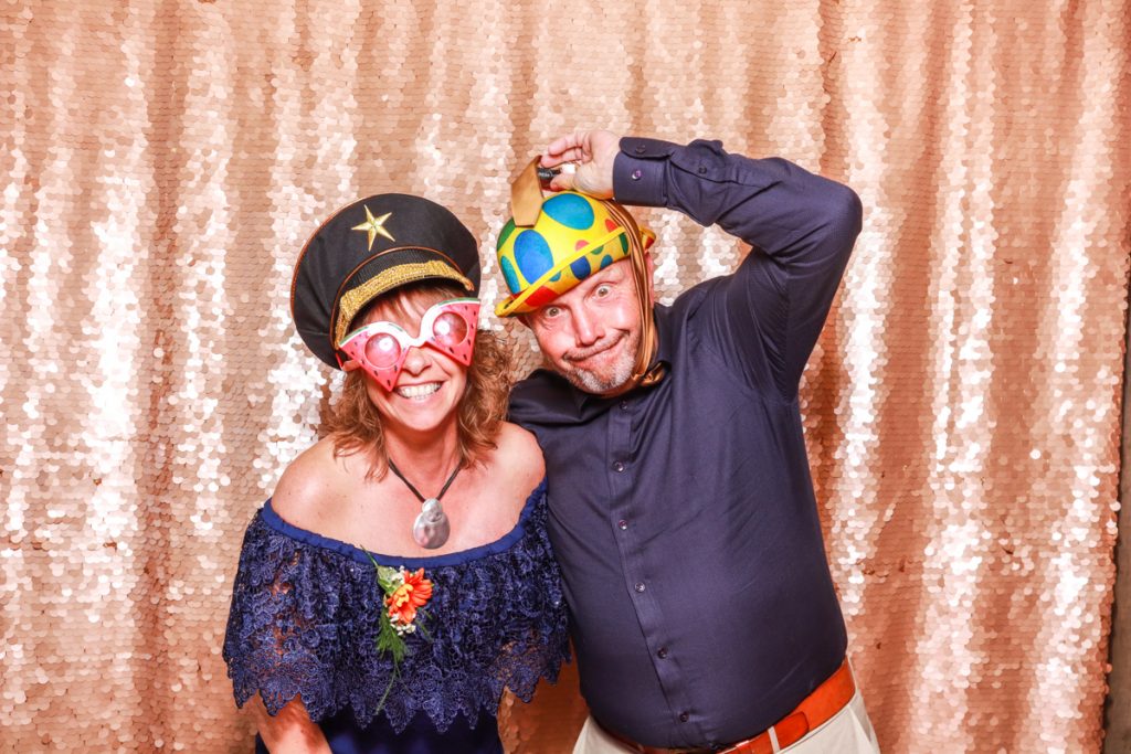 2 funny guests and wearing hats and sunglasses, posing with props, against a blush sequins backdrop, for a Wedding Photo Booth rental