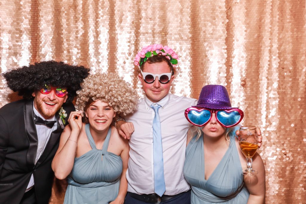 4 guests , including 2 bridesmaids in blue dresses, posing with props, against a blush sequins backdrop, for a Wedding Photo Booth rental