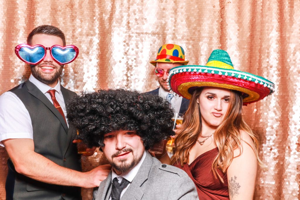 4 guests posing with props, against a blush sequins backdrop, for a Bredenbury Court Wedding Photo Booth rental