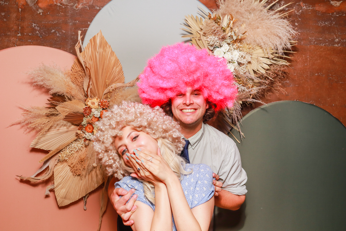 2 guests wearing fun big colourful wigs, posing against a modern arches backdrop in 3 colours, with dried flowers arrangements attached on, during a wedding venue reception party