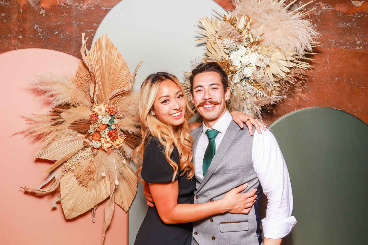 elegant couple during a wedding party at Elmore Court Gloucestershire, posing for mad hat photo booth, with a boho backdrop behind them, with floral arrangements on