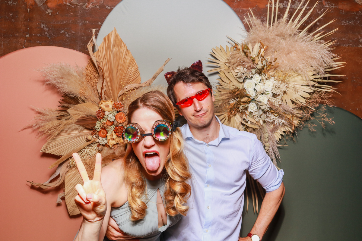 fun couple wearing props and funky sunglasses, posing against a modern arches backdrop in 3 colours, with dried flowers arrangements attached on, during gloucestershire wedding reception party