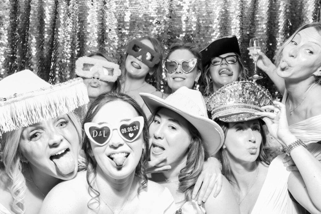 a large group of guests with the bride, posing during the wedding party entertainment, against a silver sequins backdrop, using props and hats, at The Great Tythe Barn wedding venue