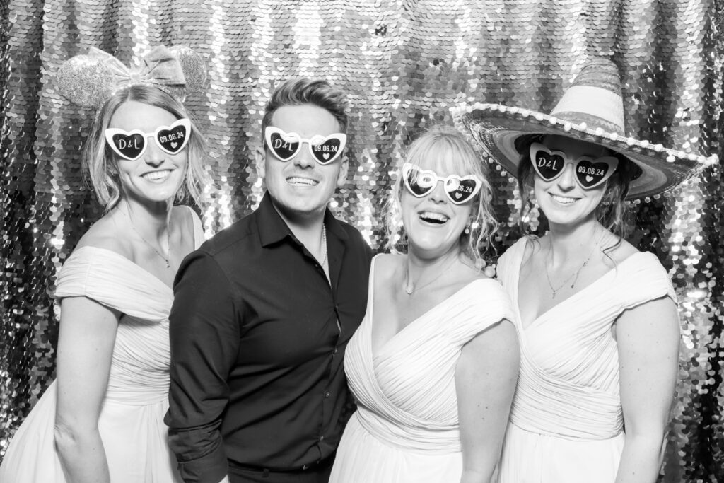 bride and groom with bridesmaids, heart shaped sunglasses during the wedding party entertainment, against a silver sequins backdrop, using props and hats, at The Great Tythe Barn wedding venue.