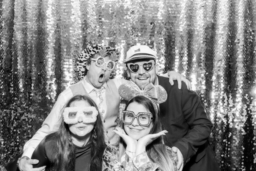 4 guests posing in silly ways, during the wedding reception entertainment, against a silver sequins backdrop, using props and hats, for mad hat photo booth rental, Cotswolds