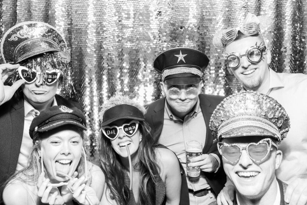 a large group of guests with the bride, posing during the wedding party entertainment, against a silver sequins backdrop, using props and hats, at The Great Tythe Barn wedding venue