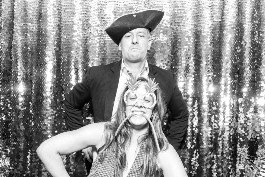 2 guests, posing during the wedding party entertainment, against a silver sequins backdrop, using props and hats, at The Great Tythe Barn wedding venue