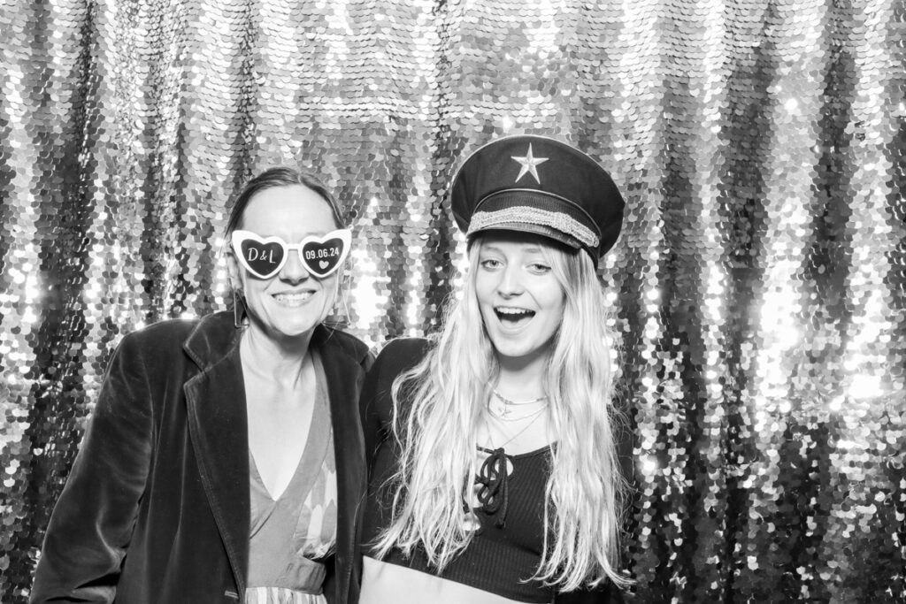 2 guests posing during the wedding party entertainment, against a silver sequins backdrop, using props and hats, at The Great Tythe Barn wedding venue