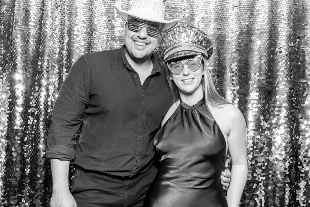 2 guests posing during the wedding party entertainment, against a silver sequins backdrop, using props and hats, during the event