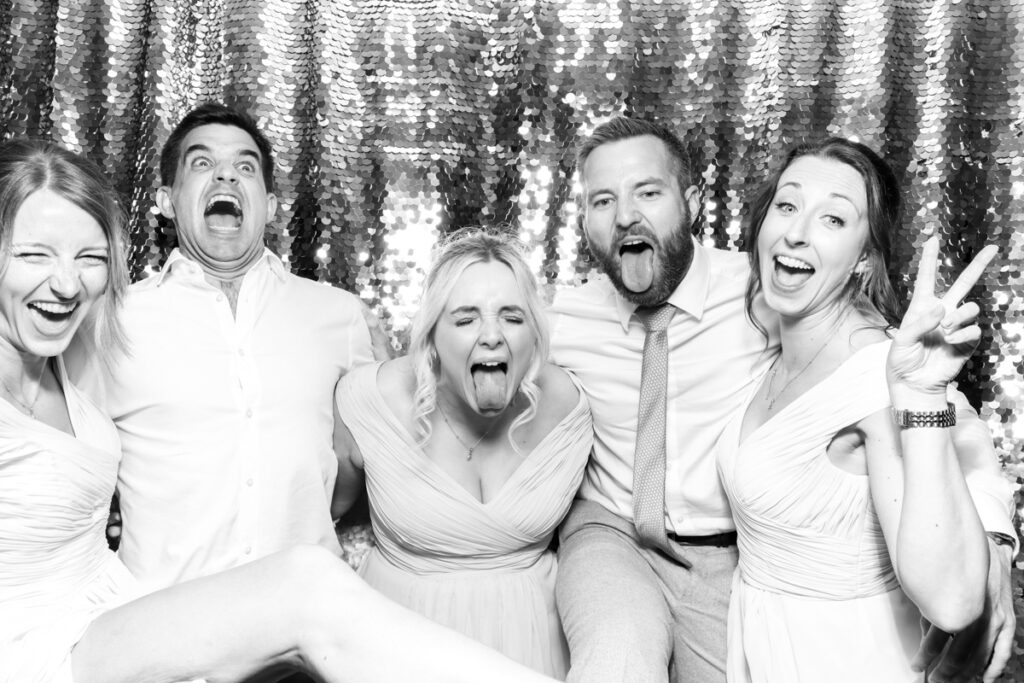 a large group of guests with the bride, posing during the wedding party entertainment, against a silver sequins backdrop, using props and hats, at The Great Tythe Barn wedding venue