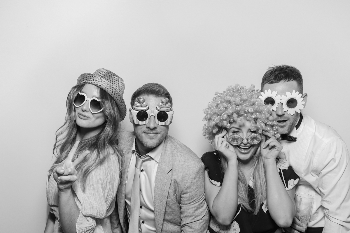 Four guests, including the bride, smiling and wearing fun sunglasses and hats in the Kardashian style photo booth B&W