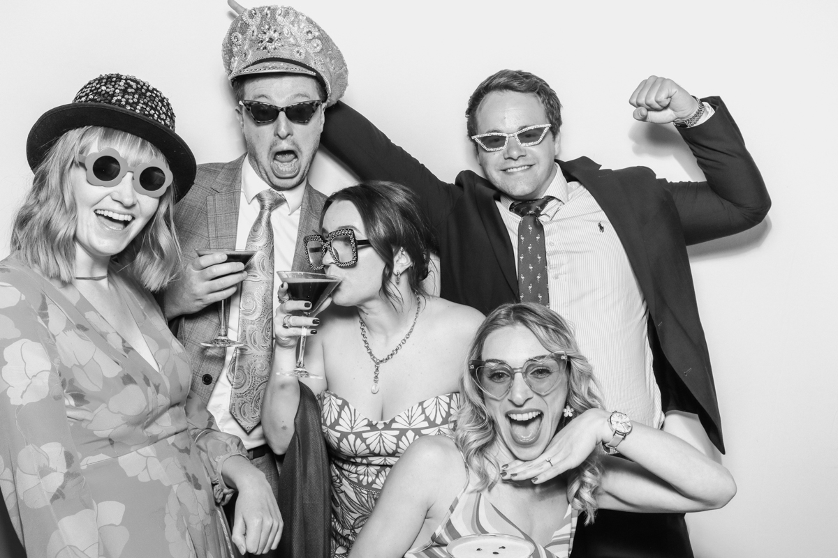 "A group of five guests posing closely together, smiling and having fun in front of the photo booth, during a wedding event