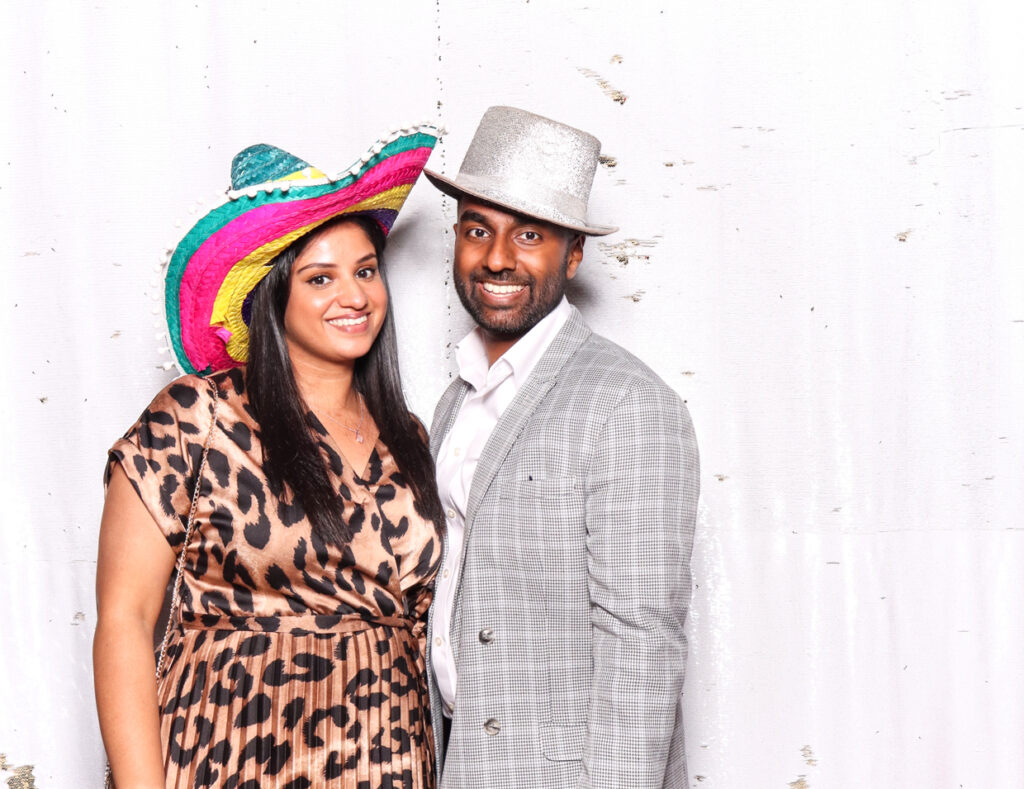 A smiling couple, the woman wearing a leopard print dress and a colourful sombrero, and the man in a checked suit with a silver glittery top hat, posing against a white backdrop during a indian wedding at sudeley castlen