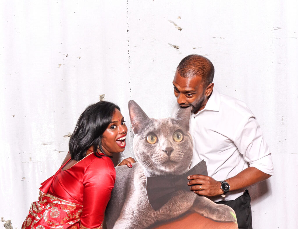 A couple enthusiastically posing with a cutout cardboard print of a cat, the woman in a red dress and the man in a white shirt, against a white backdrop during a sudeley castle wedding photo booth hire
