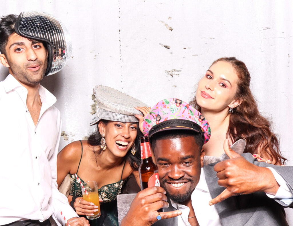 Four guests having fun, wearing colourful hats and holding drinks, one of them holding a beer bottle, posing against a white backdrop at indian-british wedding at sudeley castle