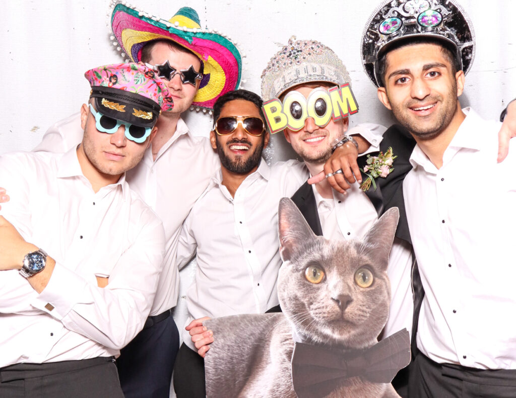 Five men wearing colourful hats and glasses, smiling and posing together with a cutout cardboard print of a cat, against a white backdrop with the photo booth rental at sudeley castle indian wedding