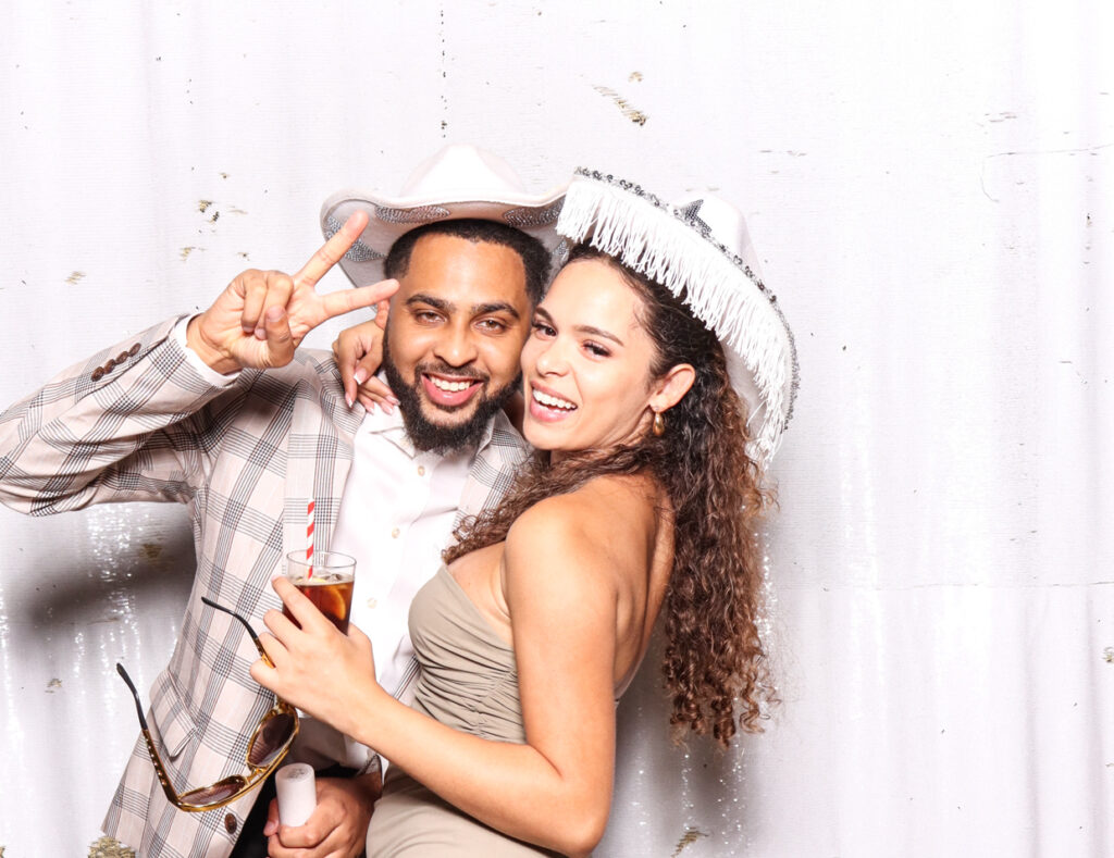 A cheerful couple, the man in a plaid suit and white cowboy hat giving a peace sign, and the woman in a light dress with a white cowboy hat, posing against a white backdrop for a sudeley castle wedding photo booth rental
