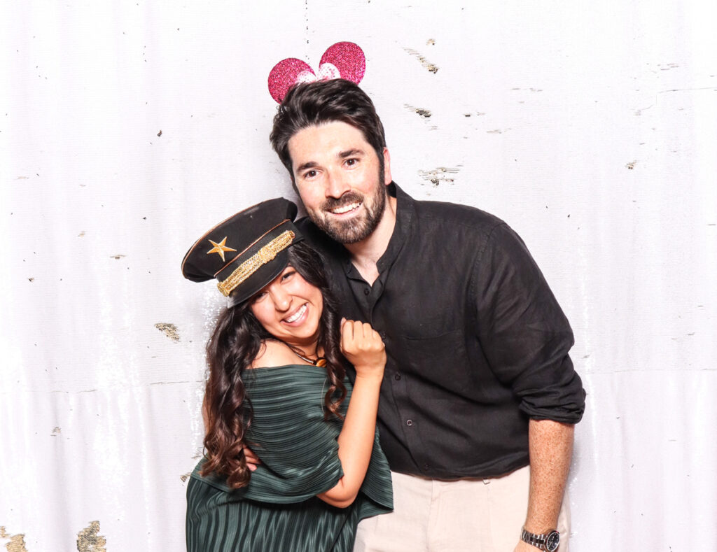 A joyful couple, the woman wearing a green dress and a captain's hat, and the man in a black shirt with heart-shaped antennae, posing against a white backdrop at an indian wedding at sudeley castle