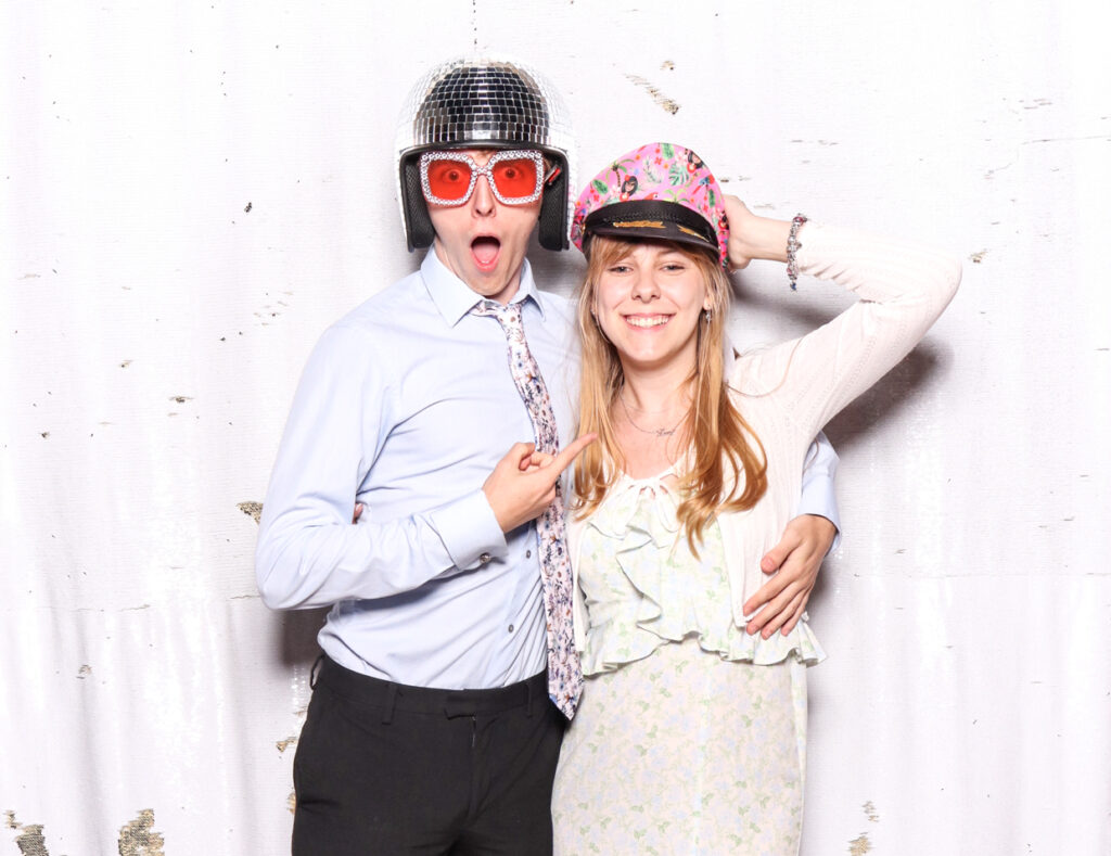 A happy couple, the man wearing a disco ball helmet and red glasses, and the woman in a floral dress with a colourful hat, posing against a white backdrop.