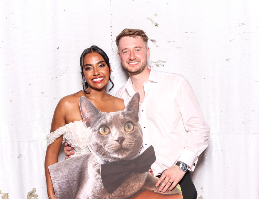 The bride and groom smiling while holding a cutout cardboard print of their cat, against a white backdrop. The bride is in a white dress and the groom in a white shirt. indian wedding at sudeley castle