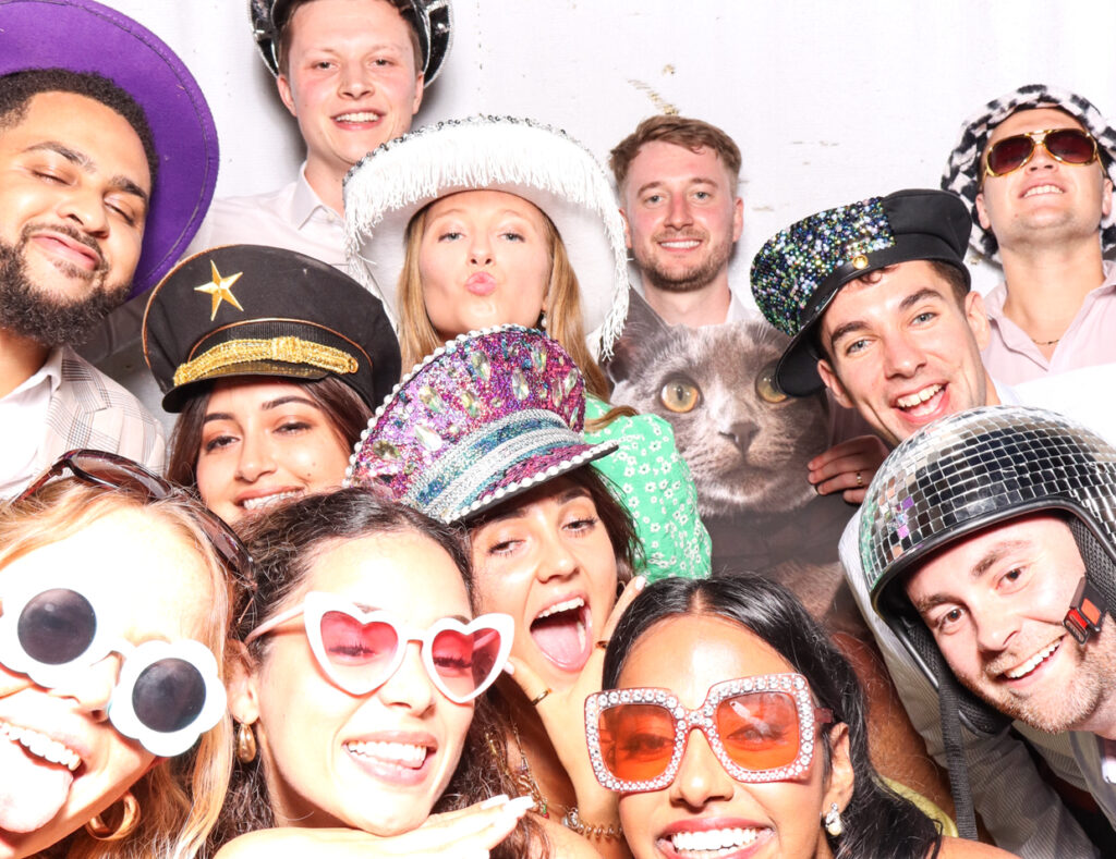 A large group of wedding guests, all wearing fun hats and glasses, smiling and posing together with a cutout cardboard print of a cat, against a white backdrop.