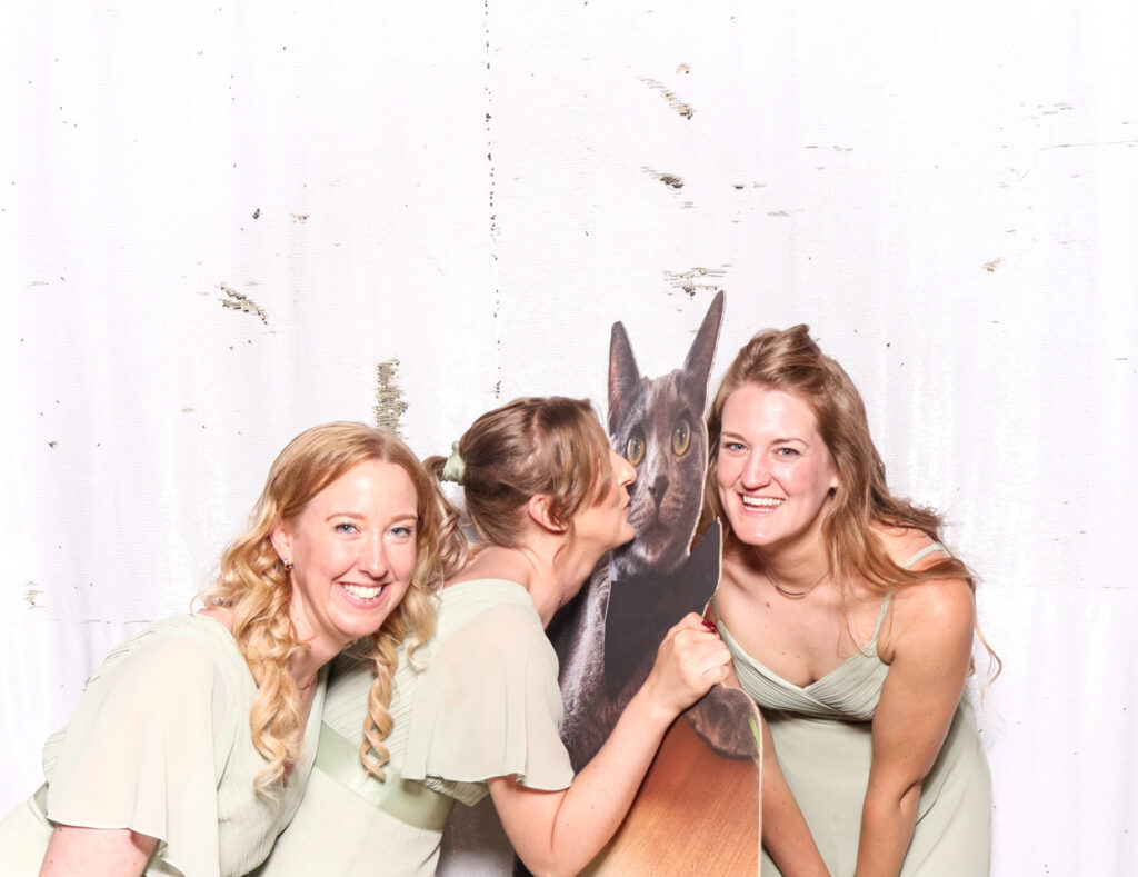 Three women, dressed in light green dresses, smiling and posing with a cutout cardboard print of a cat, against a white backdrop