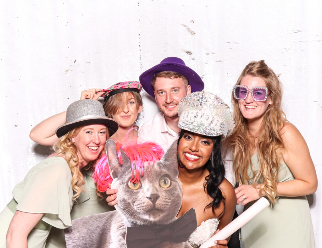 The bride, groom, and three guests, all wearing fun hats and glasses, smiling and posing together with a cutout cardboard print of a cat, against a white backdrop
