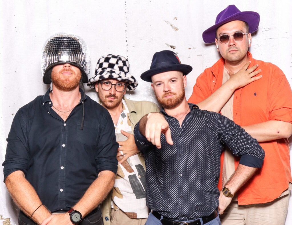 Four men posing together in fun hats and glasses, with one wearing a disco ball helmet, against a white backdrop at an indian wedding at sudeley castle 