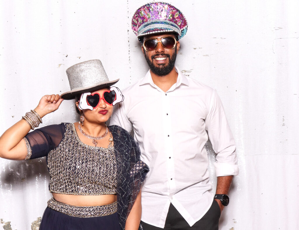 A couple, the woman dressed in a traditional Indian outfit with a silver top hat and heart-shaped glasses, and the man in a white shirt with a colourful captain's hat, posing against a white backdrop.