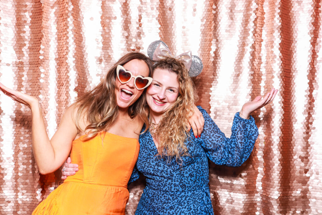 2 guests using the photo booth hire during an event, with a blush sequins backdrop and heart shaped glasses