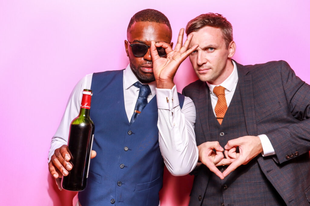 Two men in formal attire posing in front of a bubblegum pink photo booth backdrop. One man holds a bottle of wine and wears sunglasses, while the other man forms a hand gesture with his fingers, both looking playfully at the camera.