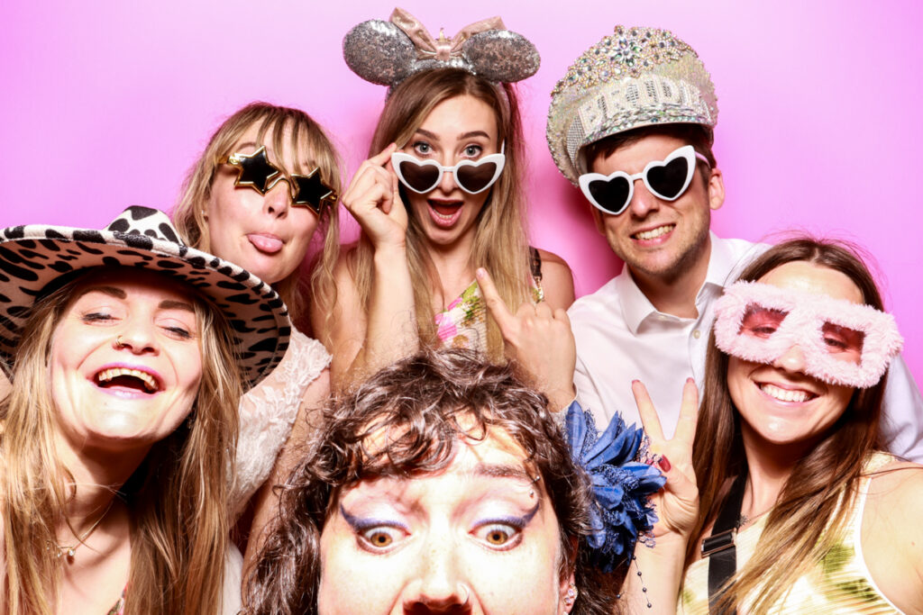  A group of six people in fun outfits and accessories, posing in front of a bubblegum pink photo booth backdrop. They are wearing pineapple-shaped glasses, star-shaped sunglasses, and leopard print hats, with one person holding a toy microphone and others making peace signs and smiling energetically.