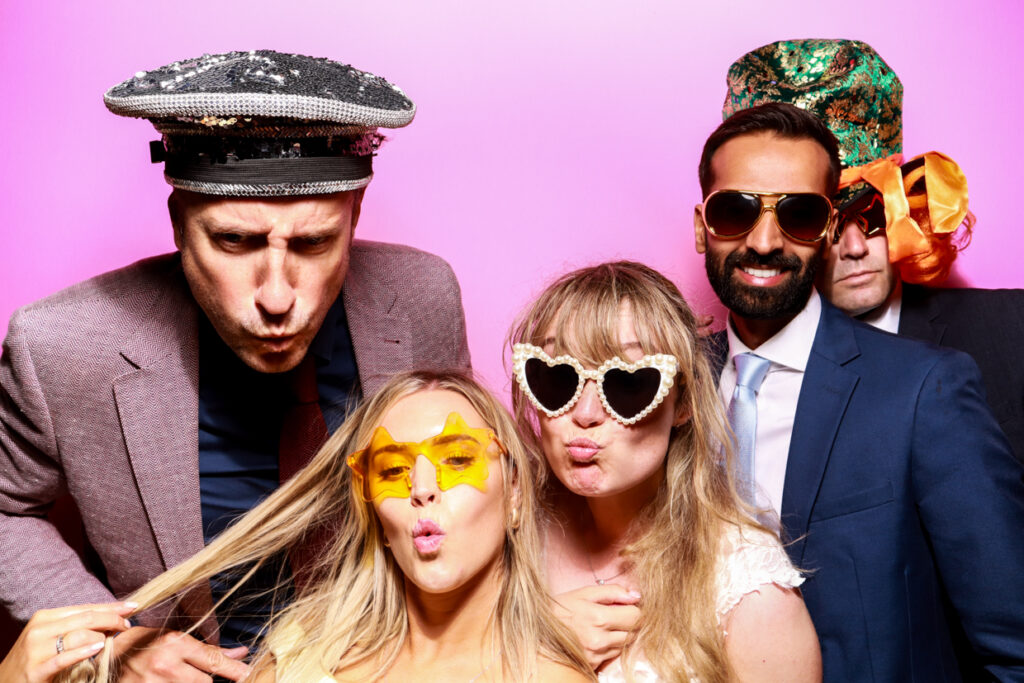 A group of five people posing together in front of a bubblegum pink photo booth backdrop. They are wearing various novelty accessories, including oversized sunglasses, sequined hats, and a large green hat with orange curls. Everyone is making playful expressions, with some pouting towards the camera.