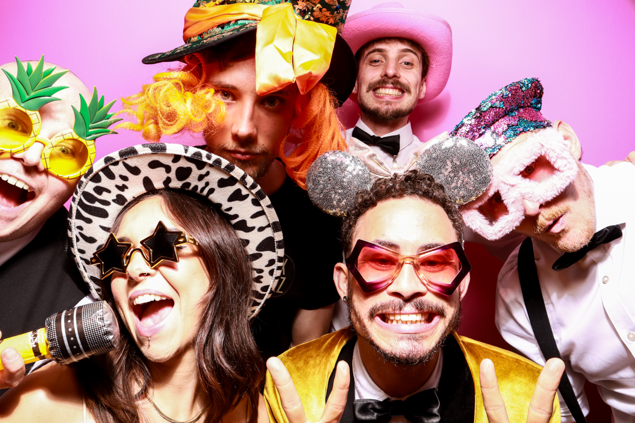 A man and a woman wearing novelty hats and glasses posing in front of a bubblegum pink photo booth backdrop. The man is wearing a tall, green hat with orange side curls, and the woman wears a leopard print hat and heart-shaped sunglasses, both making peace signs.