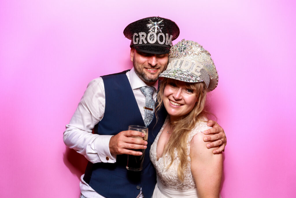 A bride and groom posing closely in front of a bubblegum pink photo booth backdrop. The groom is holding a drink and wearing a black hat labelled "Groom," while the bride is wearing a sparkling hat labelled "Bride," both smiling warmly at the camera.