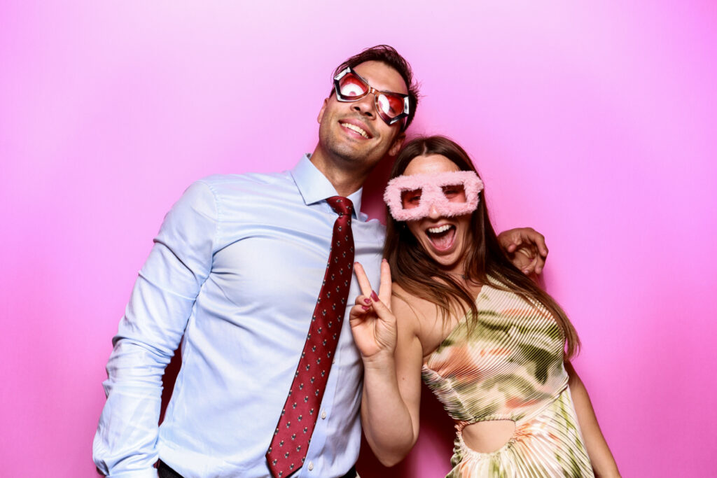 A man and woman smiling and posing in front of a bubblegum pink photo booth backdrop. The man is wearing a tie, and the woman is dressed in a patterned dress. Both are wearing oversized novelty glasses and making fun expressions.